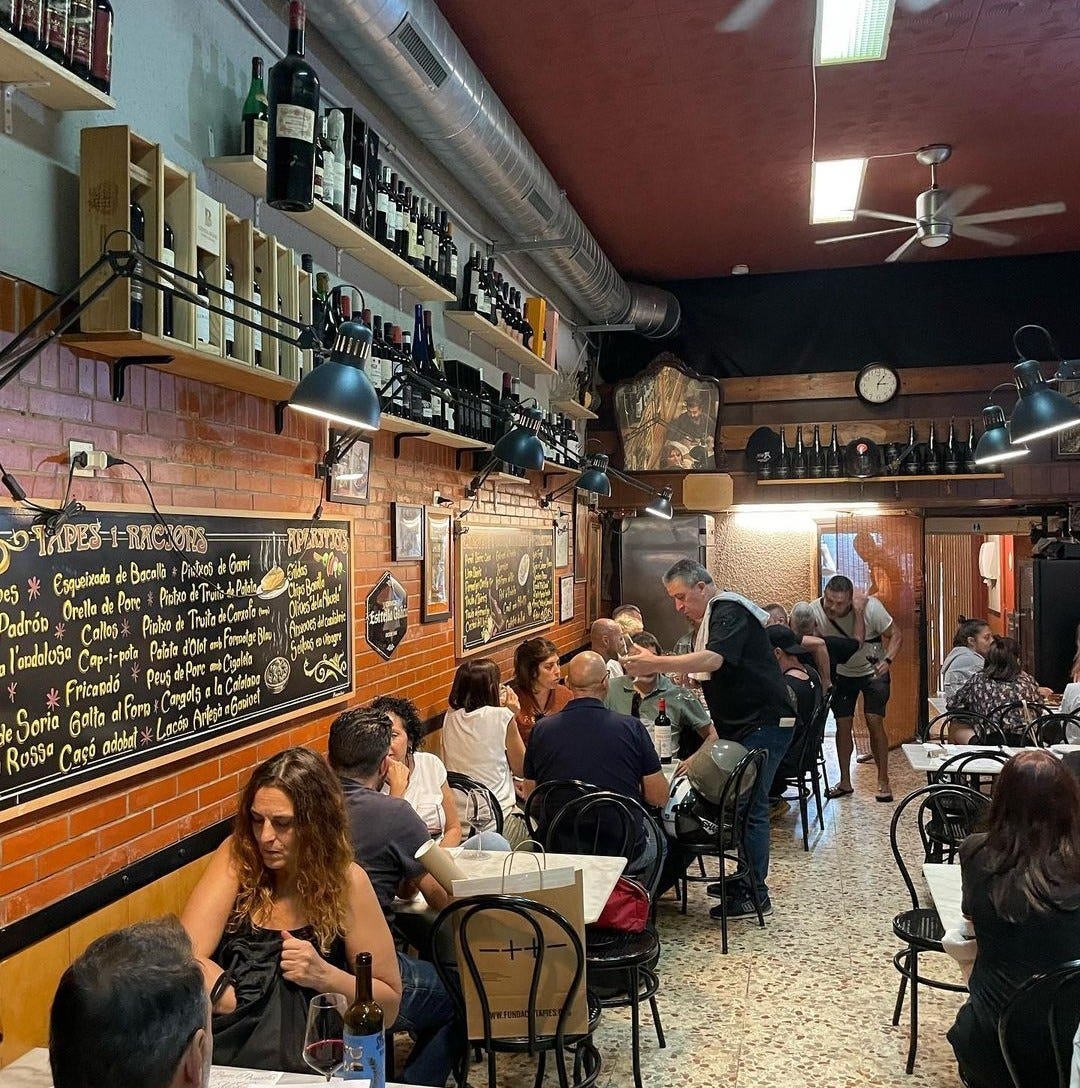 Busy dining room; 'Tapas i racions' chalkboard readable (esqueixada, callos, cap-i-pota, fricandó...)